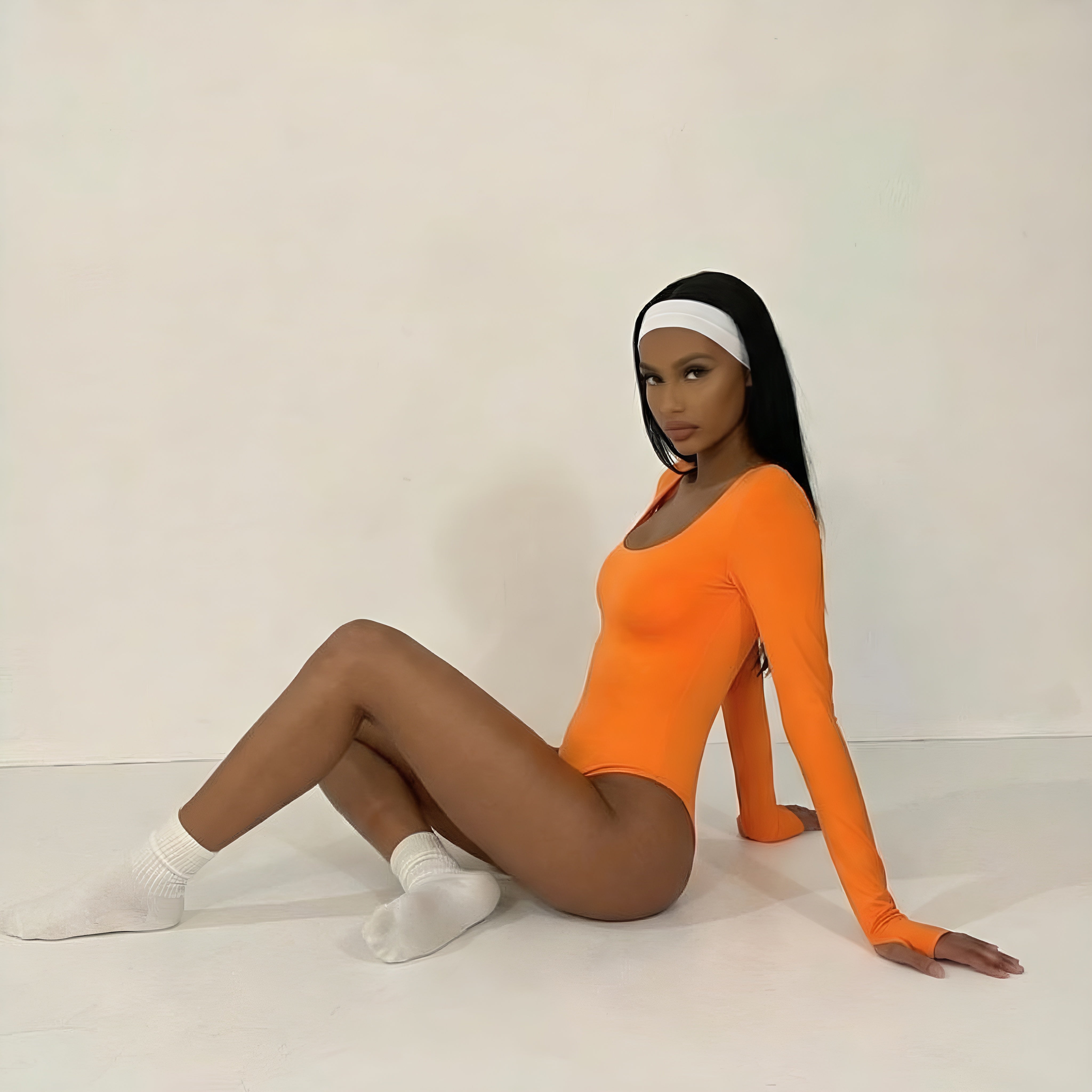 Woman wearing an orange bodysuit sitting on a white floor against a white wall. Woman shapewear bodysuit lyvia.