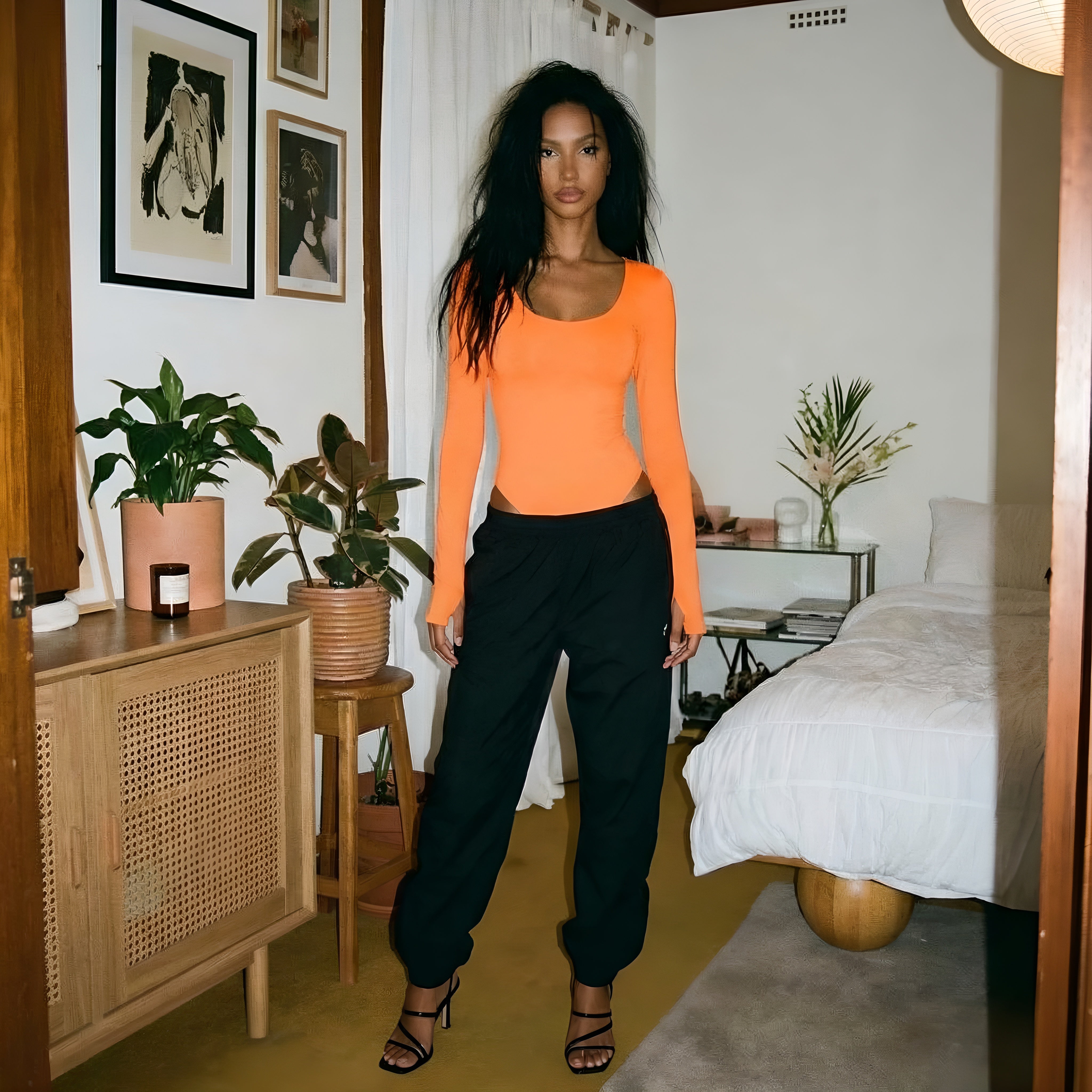 Woman in an orange top and black pants standing in a bedroom with plants and furniture. Woman shapewear orange bodysuit photoshoot lyvia.