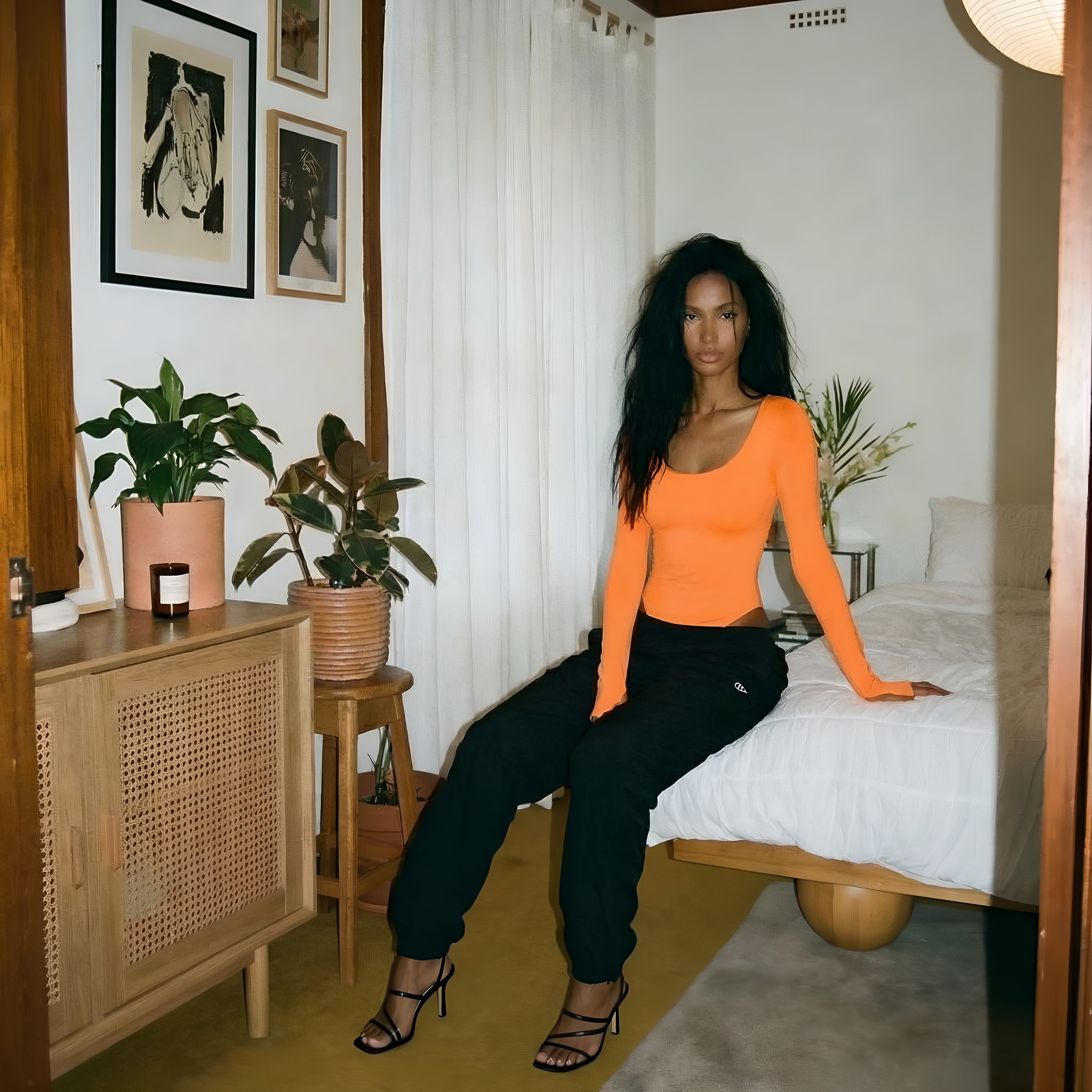 Woman in an orange top and black pants sitting on a bed in a room with plants and furniture. Orange womans bodysuit lyvia.