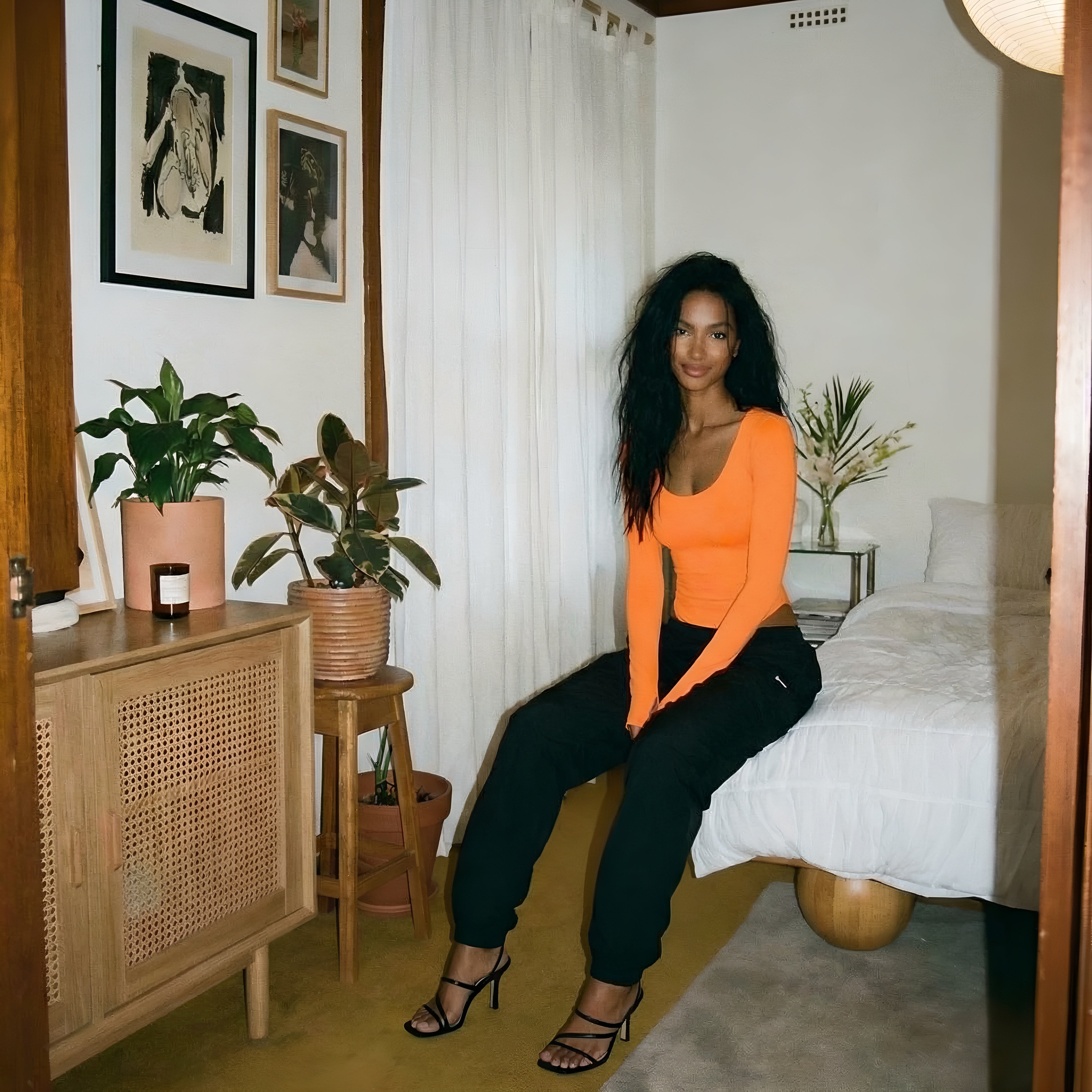 Woman in an orange top and black pants sitting in a room with plants and furniture. Orange womans bodysuit shapewear lyvia.