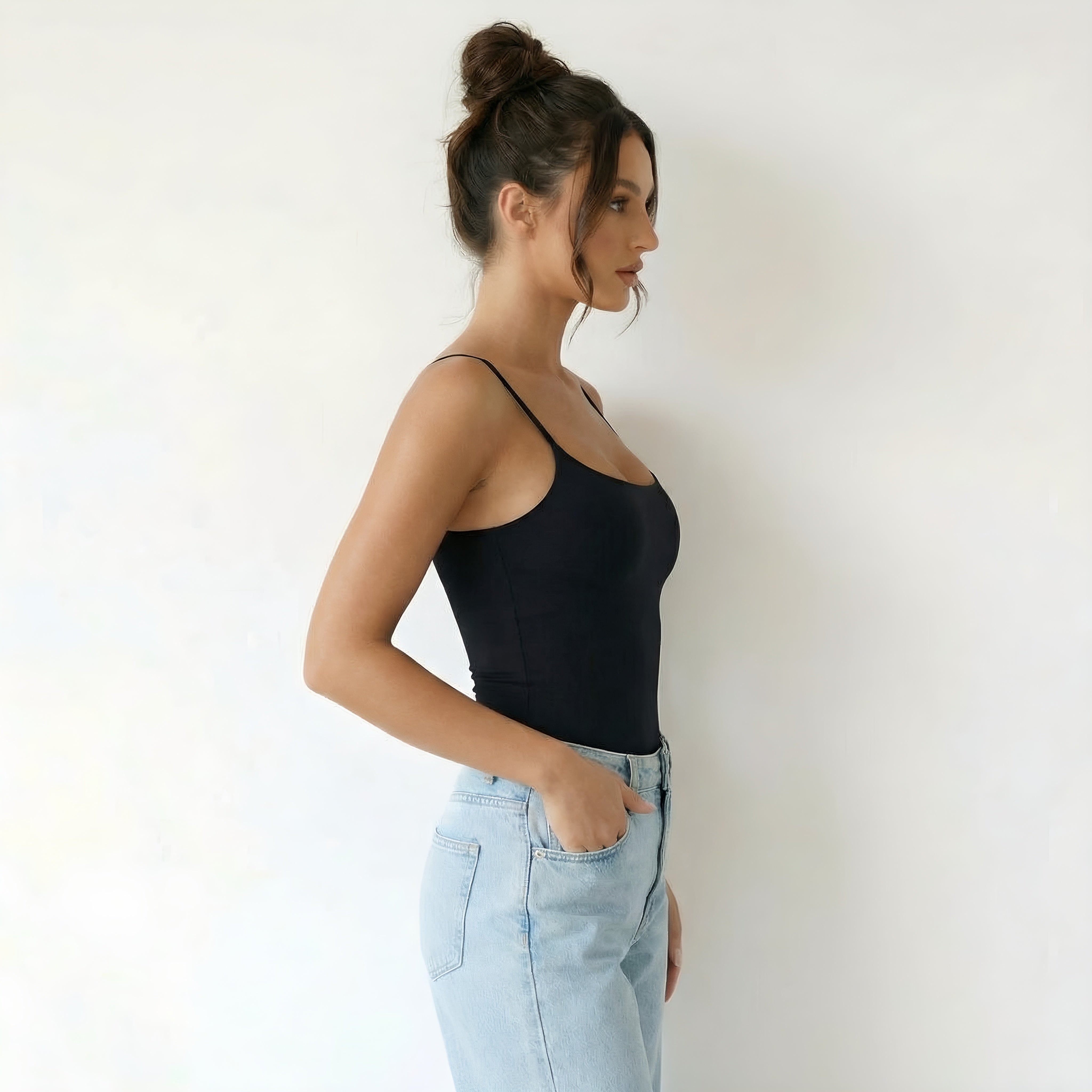 Woman wearing a black tank top and light blue jeans against a white background. Woman wearing black shapewear lyvia top