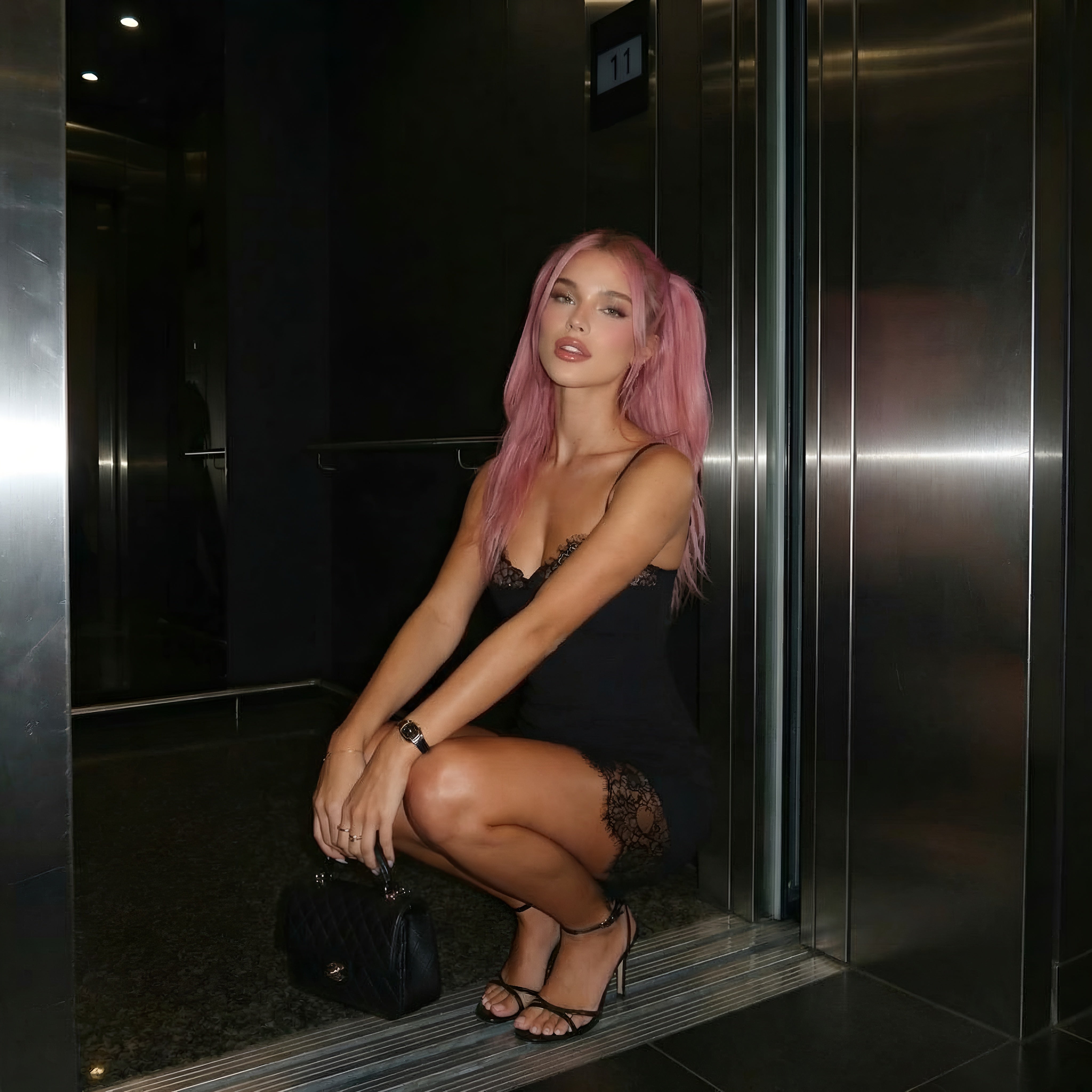 Woman with pink hair sitting inside an elevator wearing a black dress. LYVIA Midnight Lace Mini Dress