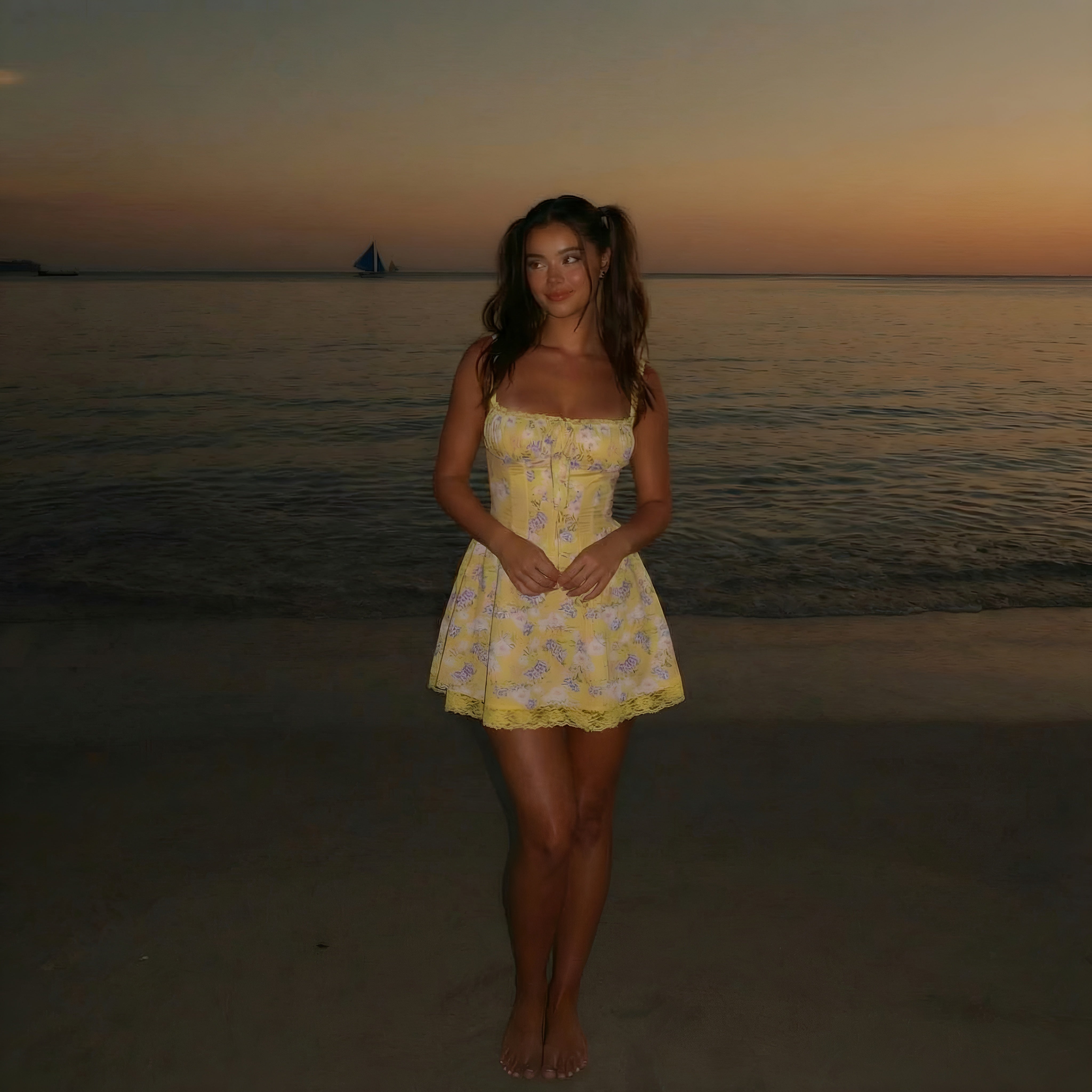Woman in a yellow dress standing on a beach at sunset with a sailboat in the distance. LYVIA Yellow Floral Meadow Bloom Mini Dress.
