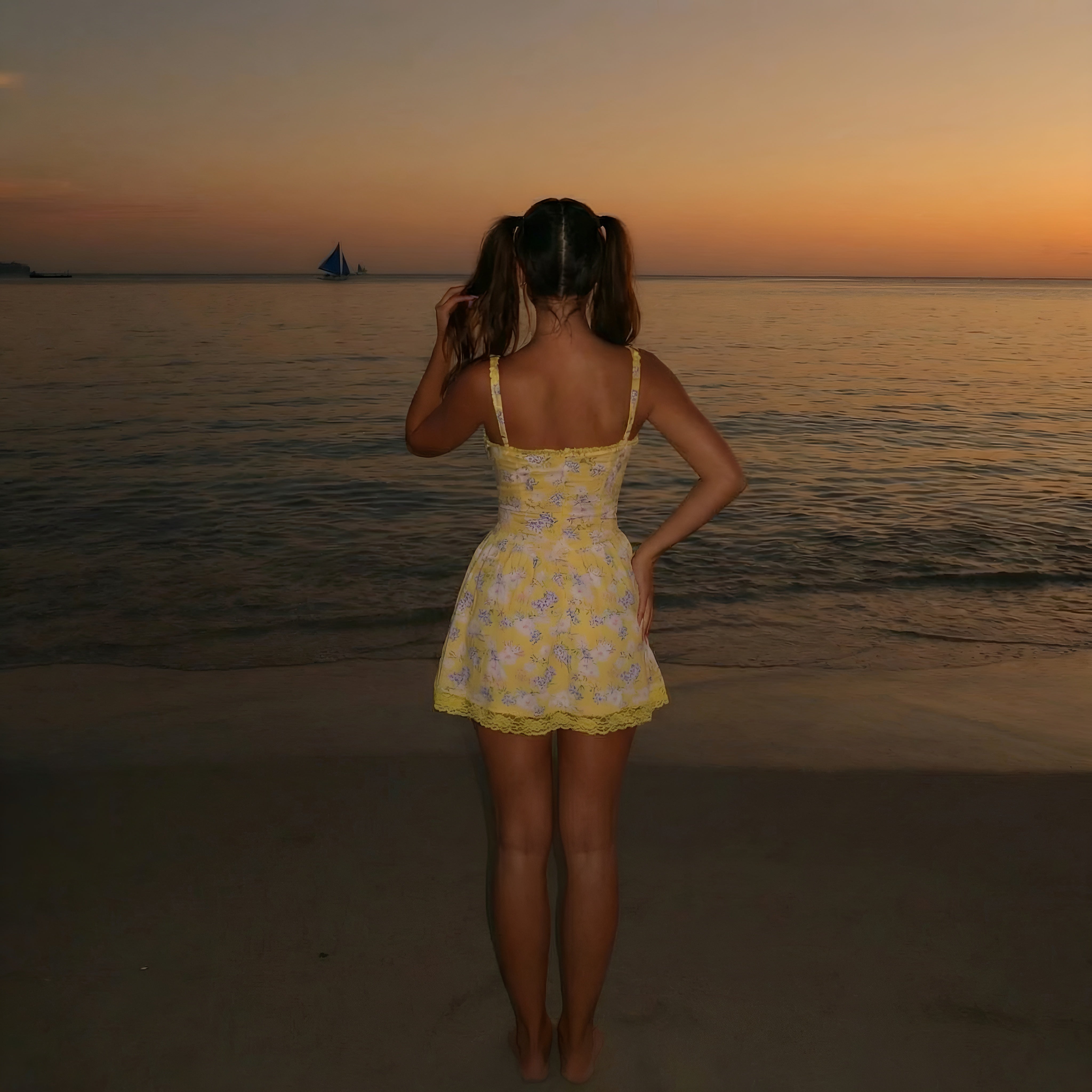 Woman in a yellow dress standing on a beach at sunset, taking a photo of a sailboat. LYVIA Yellow Floral Meadow Bloom Mini Dress.